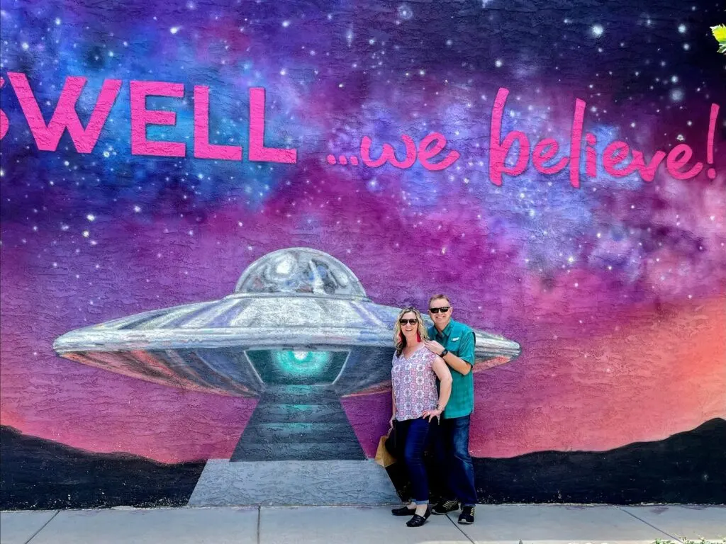 couple standing in front of colorful mural in Roswell that says "Roswell we believe" and has a space ship on it